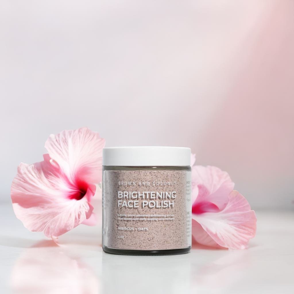 Brown and Coconut Brightening Face Polish on a glossy surface in front of pink hibiscus flowers