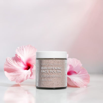 Brown and Coconut Brightening Face Polish on a glossy surface in front of pink hibiscus flowers